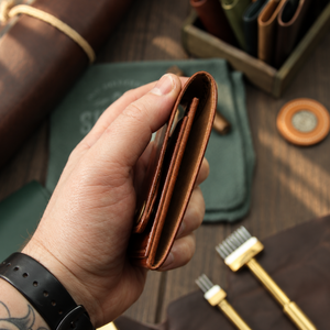 A hand holds a brown leather bifold wallet with card slots, shown in a styled workshop setting with leather tools and brushes on a wooden table.