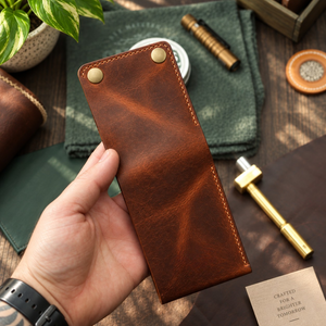 A hand holds a brown leather card holder or sleeve with two brass snap buttons, set against a styled desk background with green cloth, pens, and a plant.