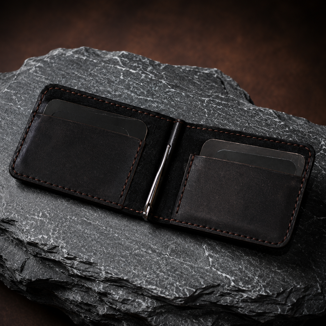 A dark brown leather money clip wallet with visible contrast stitching, shown open and resting on a textured dark gray rock.