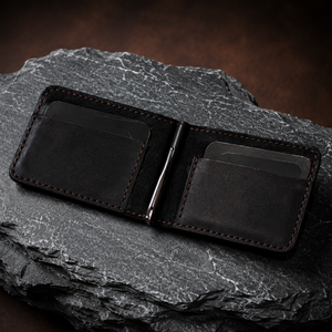 A dark brown leather money clip wallet with visible contrast stitching, shown open and resting on a textured dark gray rock.